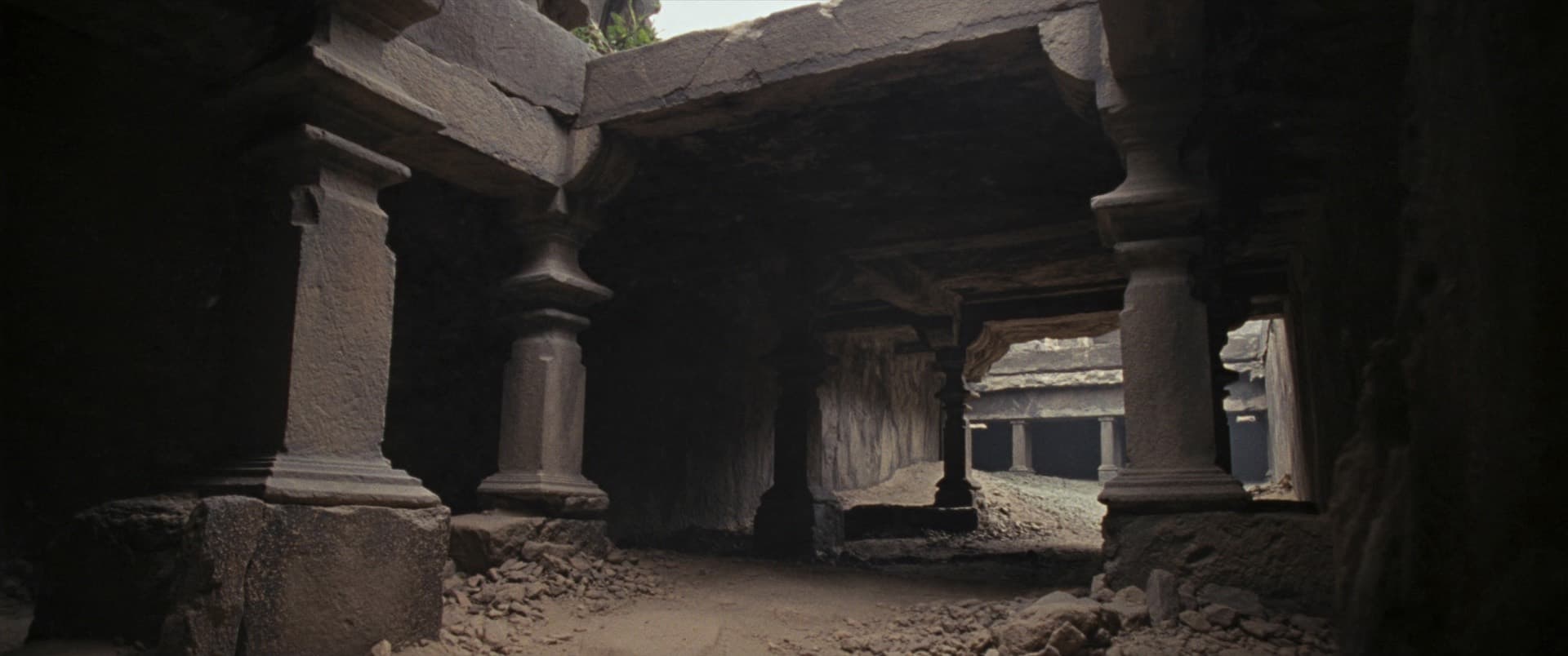 A pillared stone hall with daylight entering from one side. The far corners have gone to shadow while the near columns still hold their carving.