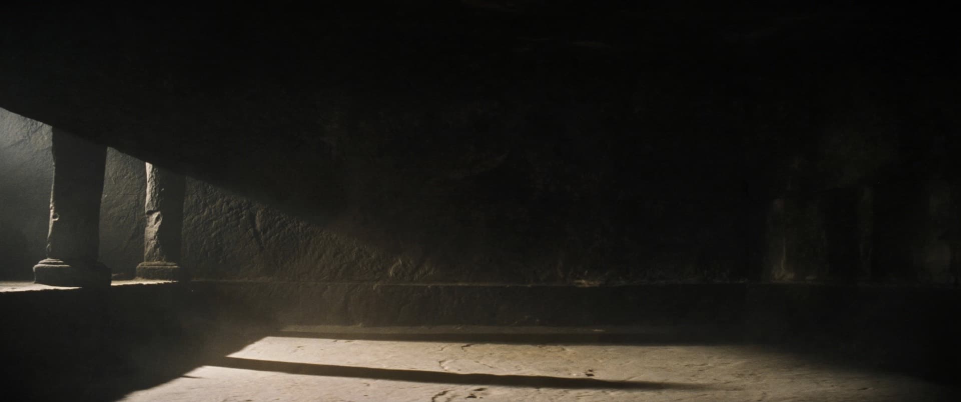 A dark hall with one low shaft of light crossing the floor. Two columns stand at the edge of it; everything beyond them is unlit stone.